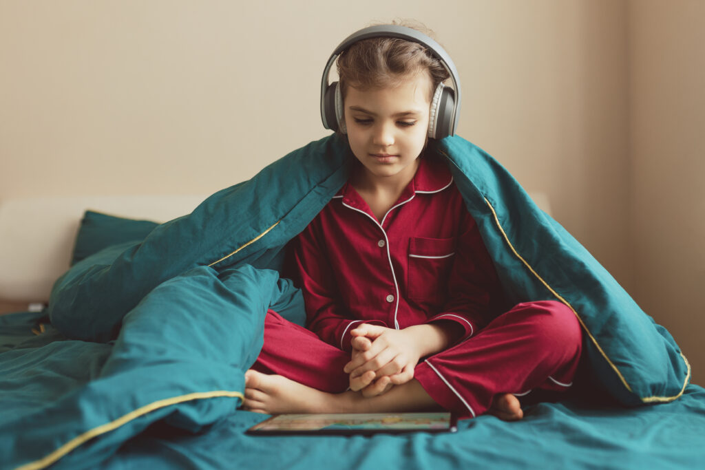 A preteen girl listening to an audiobook, wearing headphones sitting on a bed.