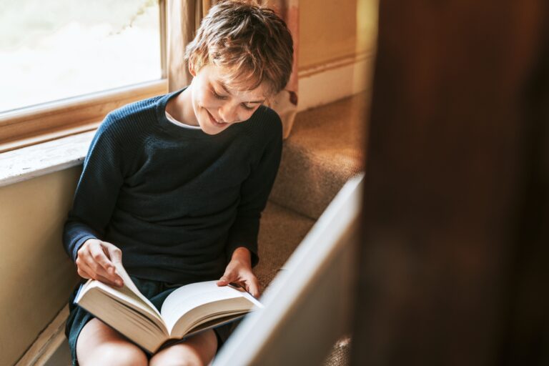 Young boy happily reading a chapter book by a window.