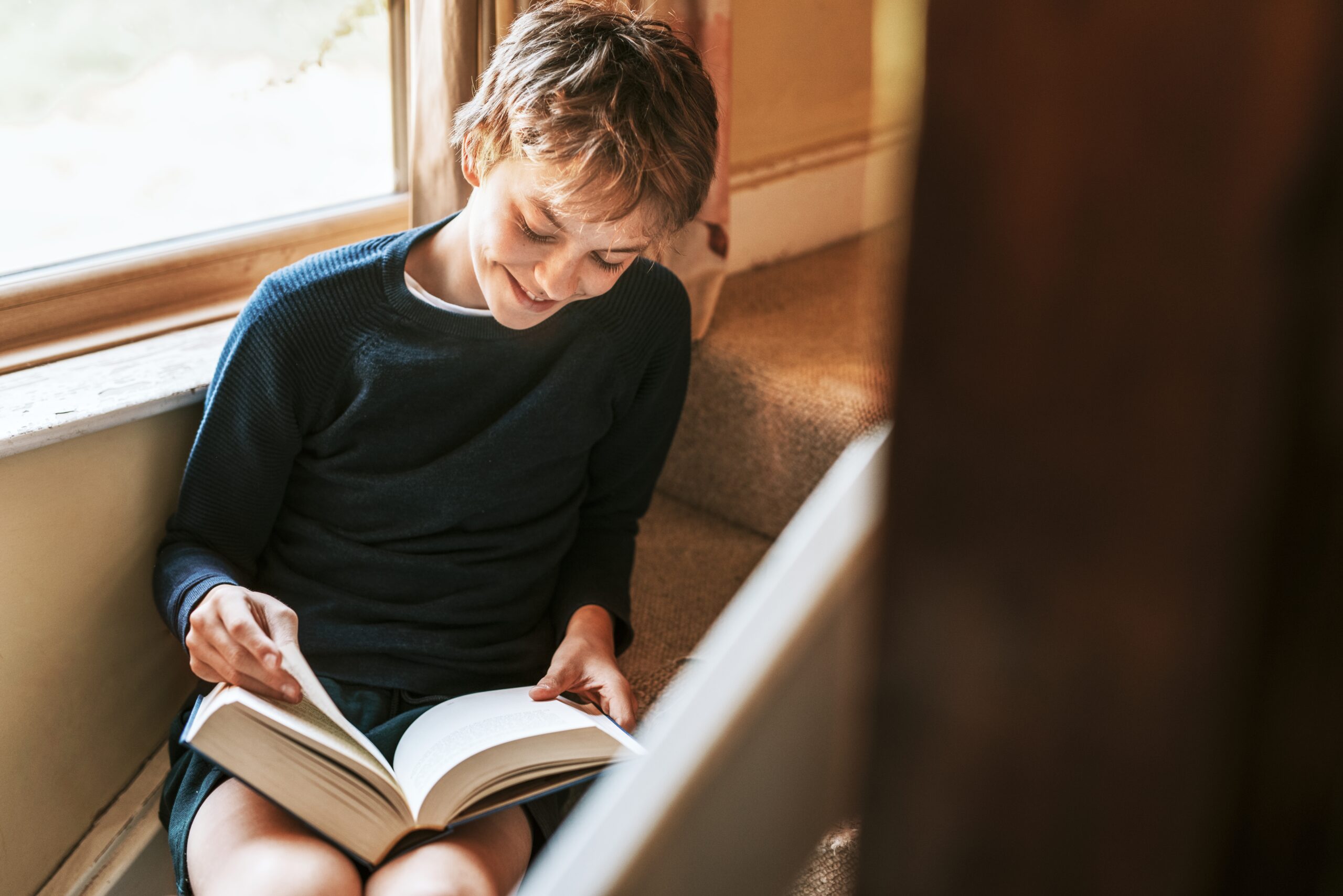 Young boy happily reading a chapter book by a window.