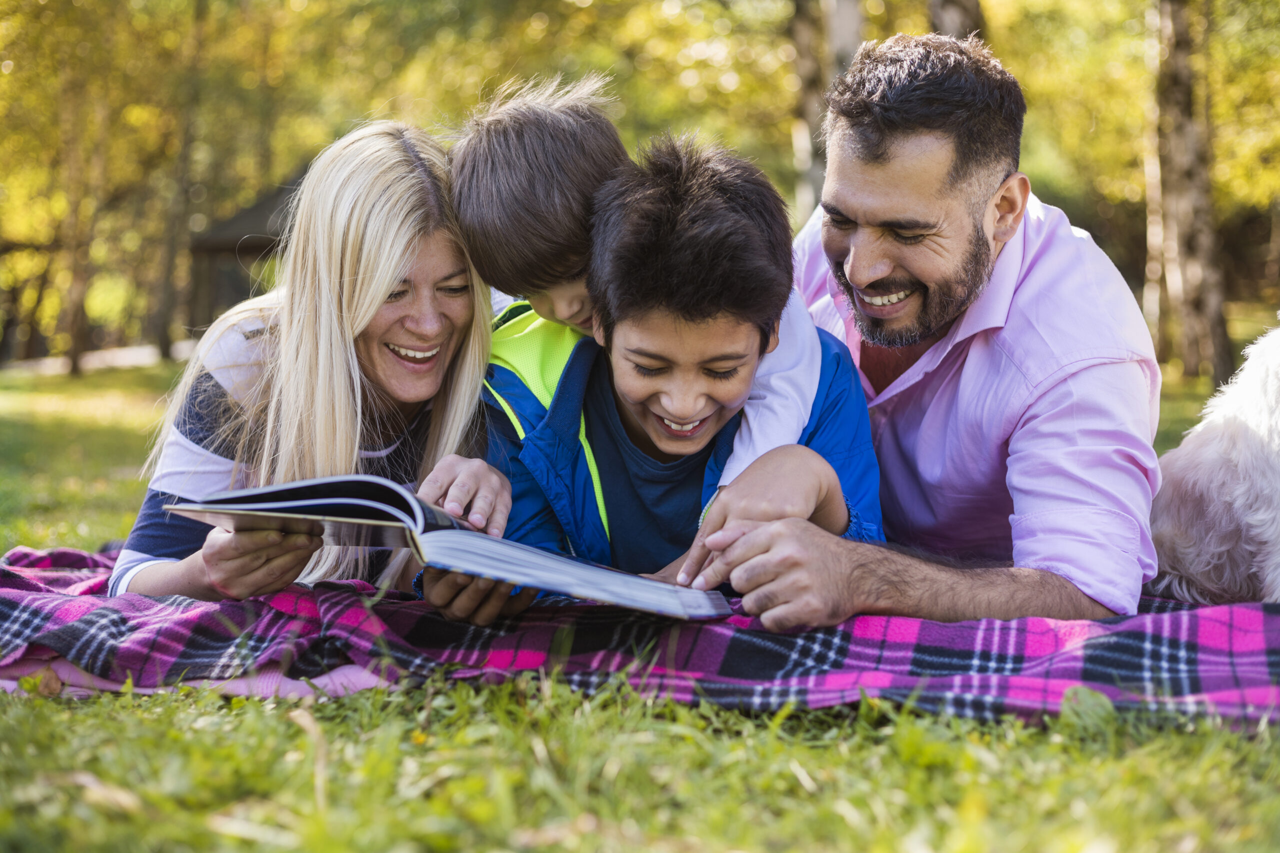 Family lying together on a blanket outdoors, smiling and laughing while sharing a book.