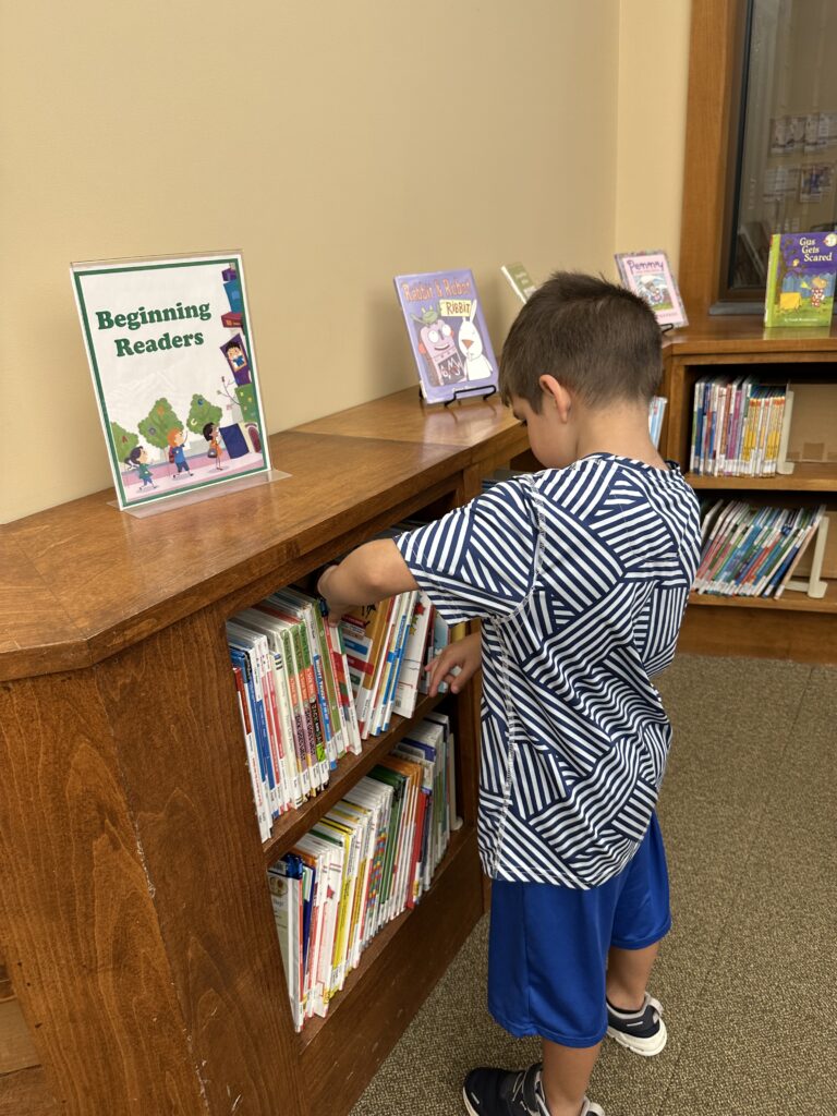 Young boy browsing the “Beginning Readers” section at the library, carefully pulling out a book while exploring early reading options.