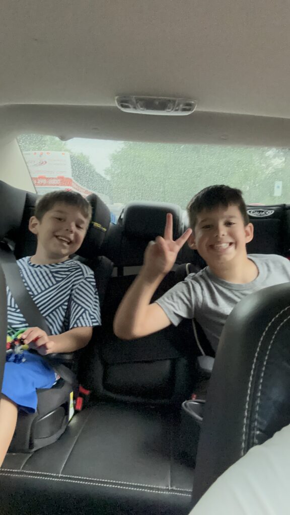 Two smiling boys sitting in the backseat after a library visit, one flashing a peace sign, showing joy and success after a trip with no tears or frustration.