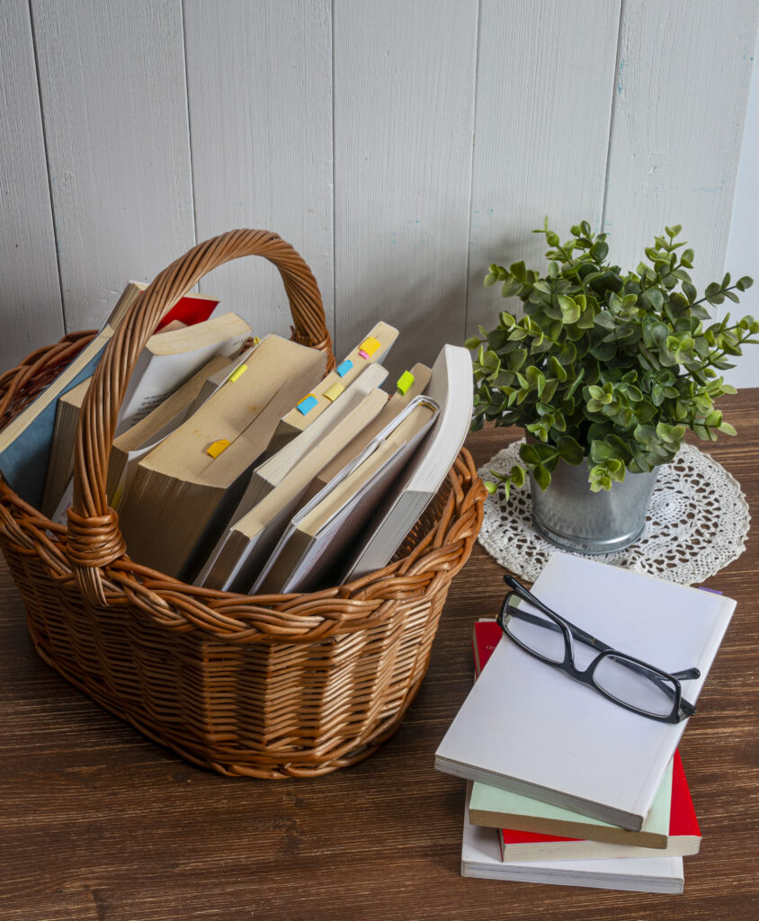 A wicker basket with some books from the library on a wooden table in the living room ready to be read. Some with favorite pages already marked so they know where to pick up from.