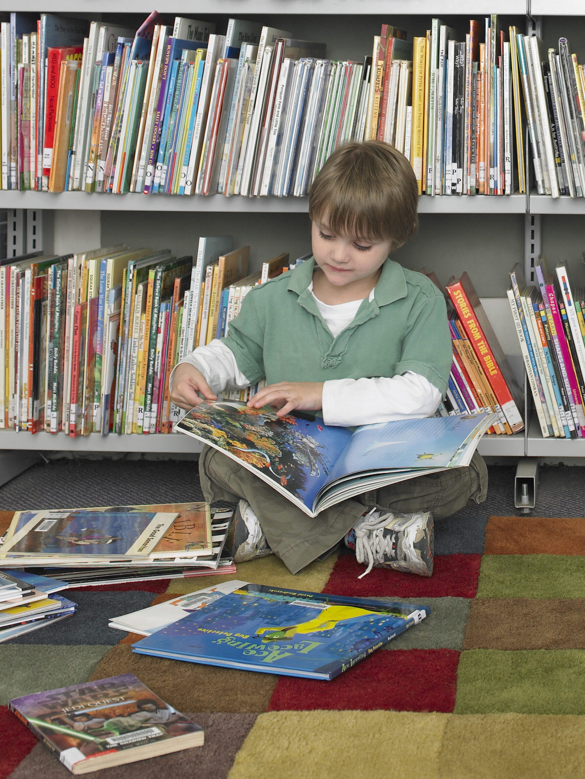 Young boy sitting the library, flipping through a book with a messy pile of books scattered around him.