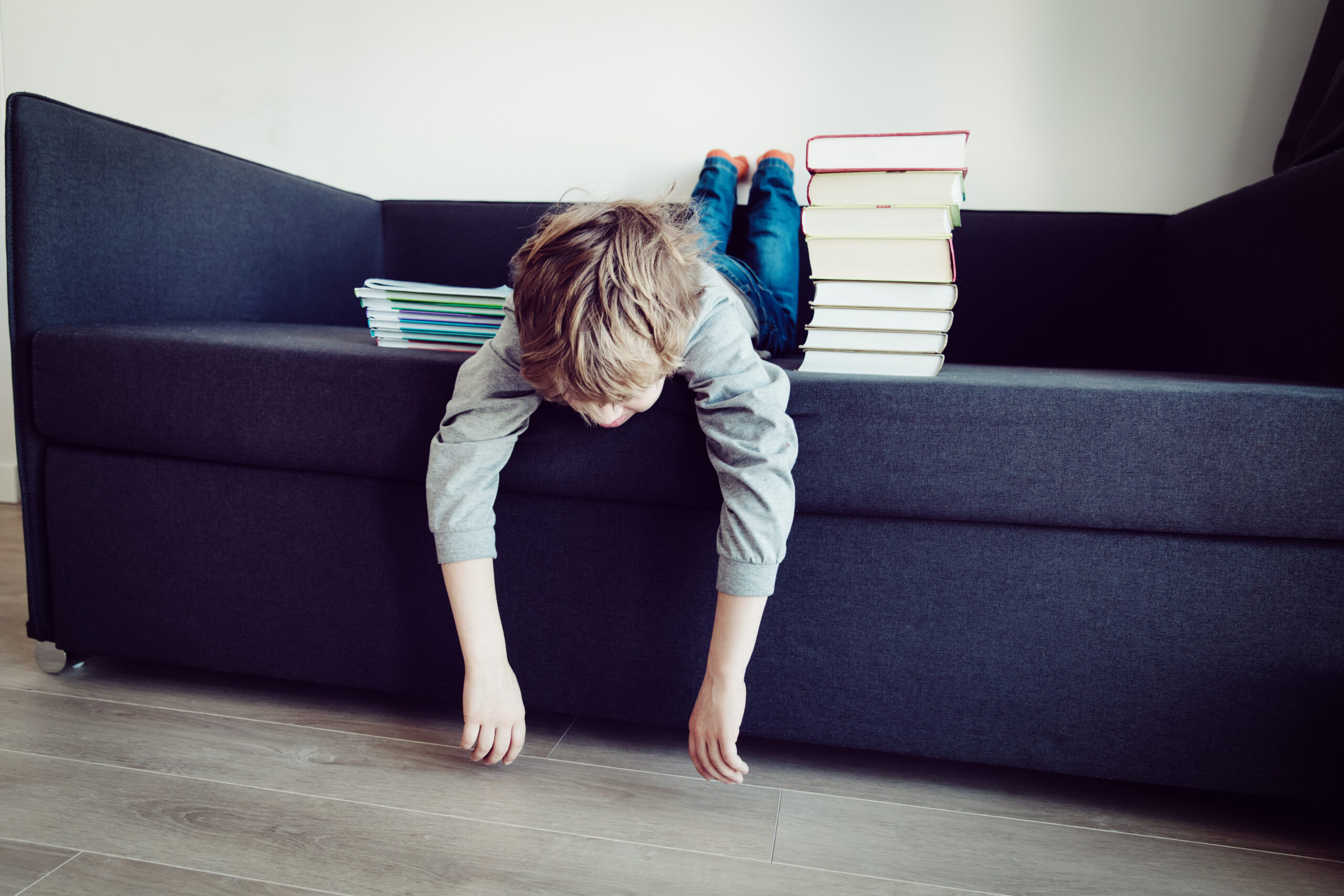 Tired elementary-aged boy lying on a couch, looking exhausted with a stack of books beside him, not reading — symbolizing back-to-school fatigue and resistance to reading.