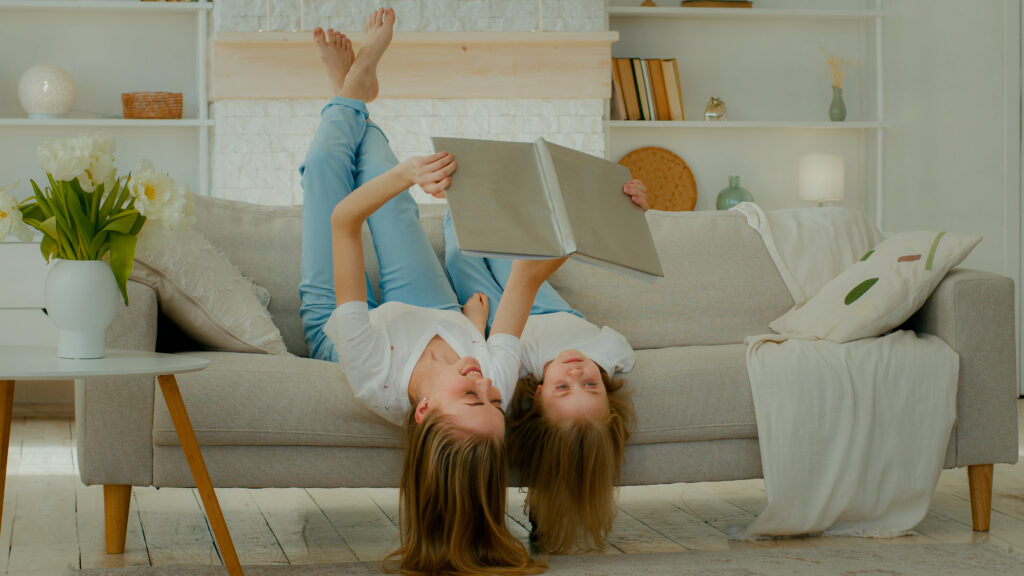 Mother and daughter lying upside down on a couch together, holding a book above their heads and laughing.