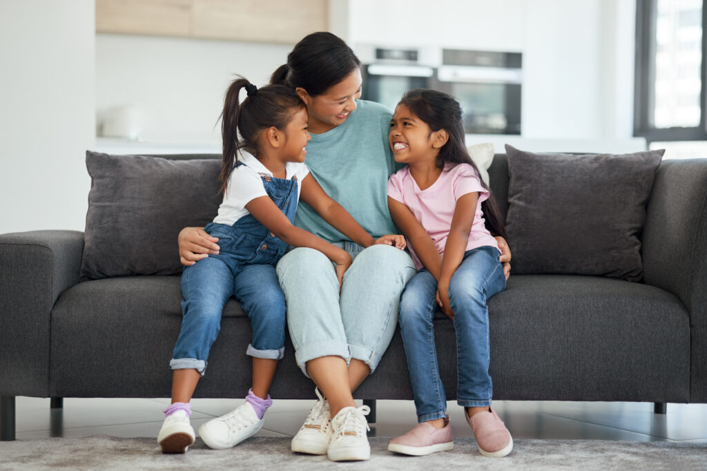 Parent sitting on the couch with two young daughters, smiling and talking together, preparing for their next library visit.